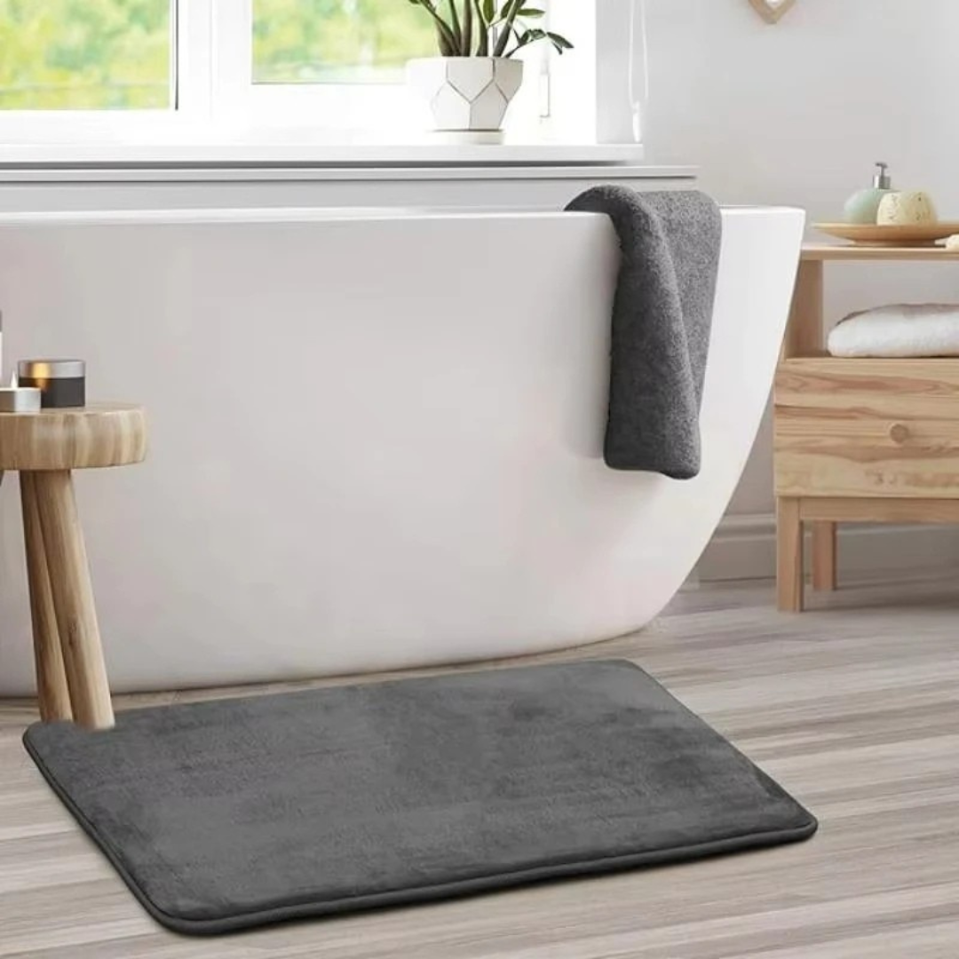 Gray bath mat in front of a bathtub with a towel draped over the edge.